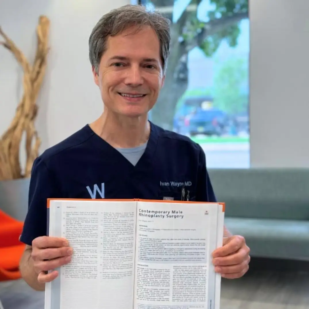 Smiling doctor in scrubs holds a journal open to rhinoplasty article.
