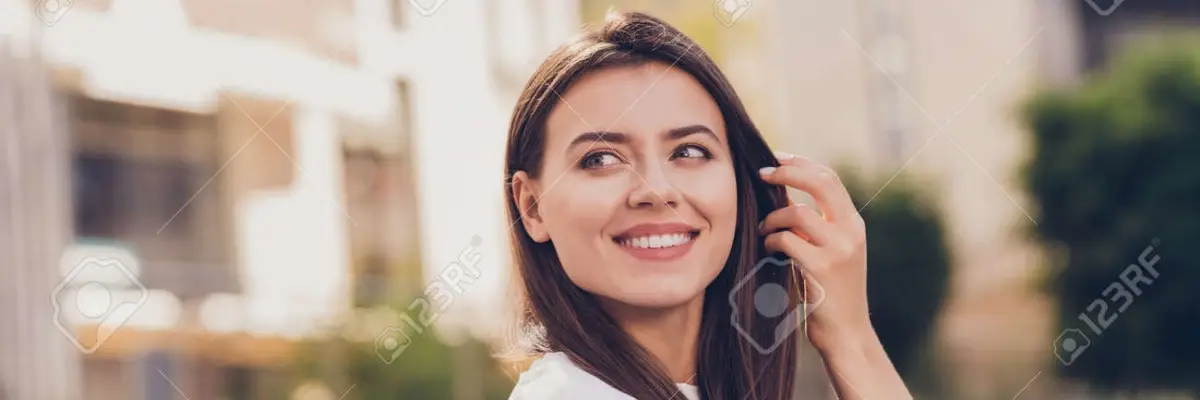 Woman with long brown hair smiles and looks to the side outdoors on a sunny day.