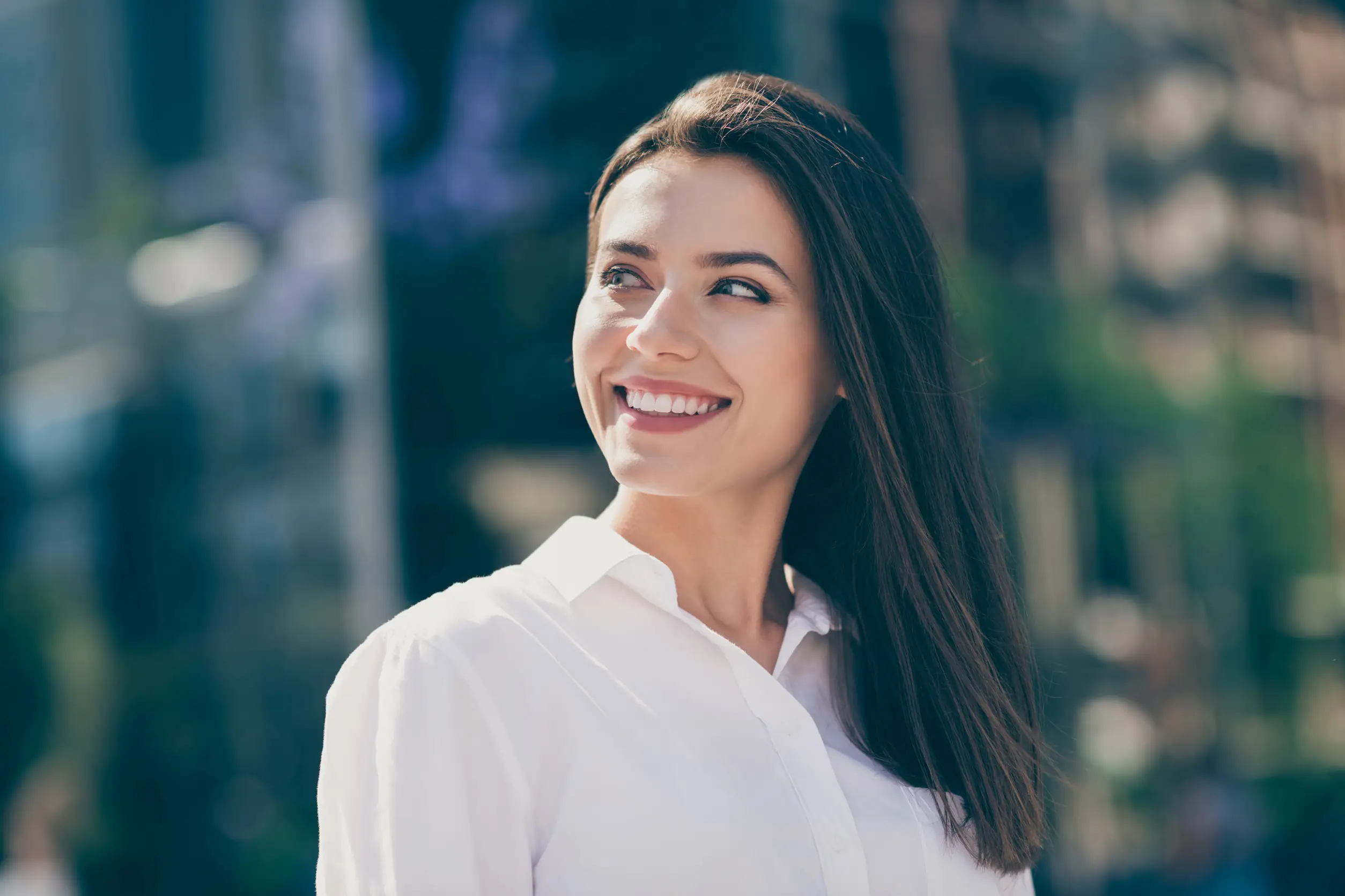 Woman with long brown hair in a white shirt smiling outdoors on a sunny day.