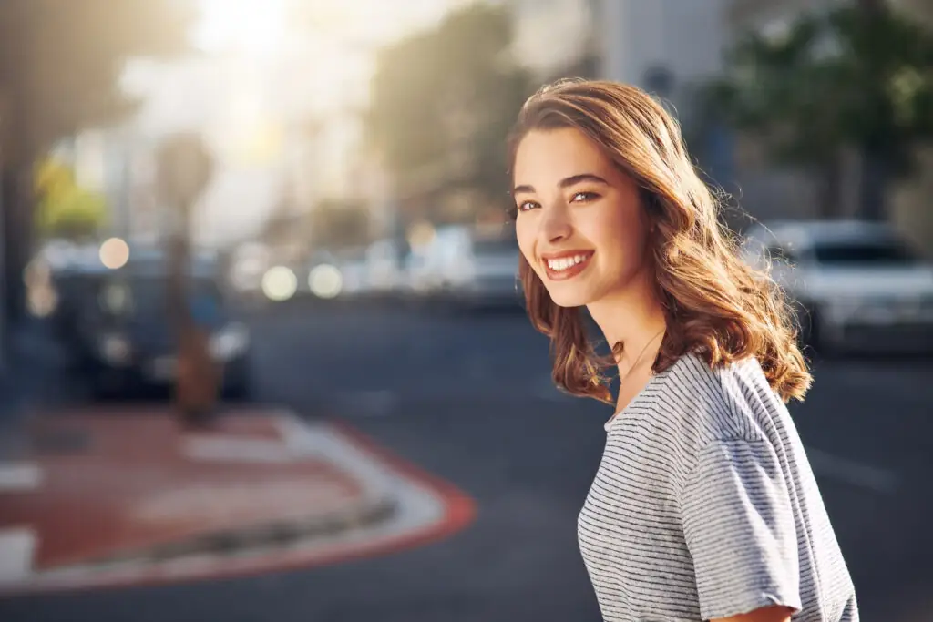 Young woman with brown hair smiles confidently on a sunny city street.