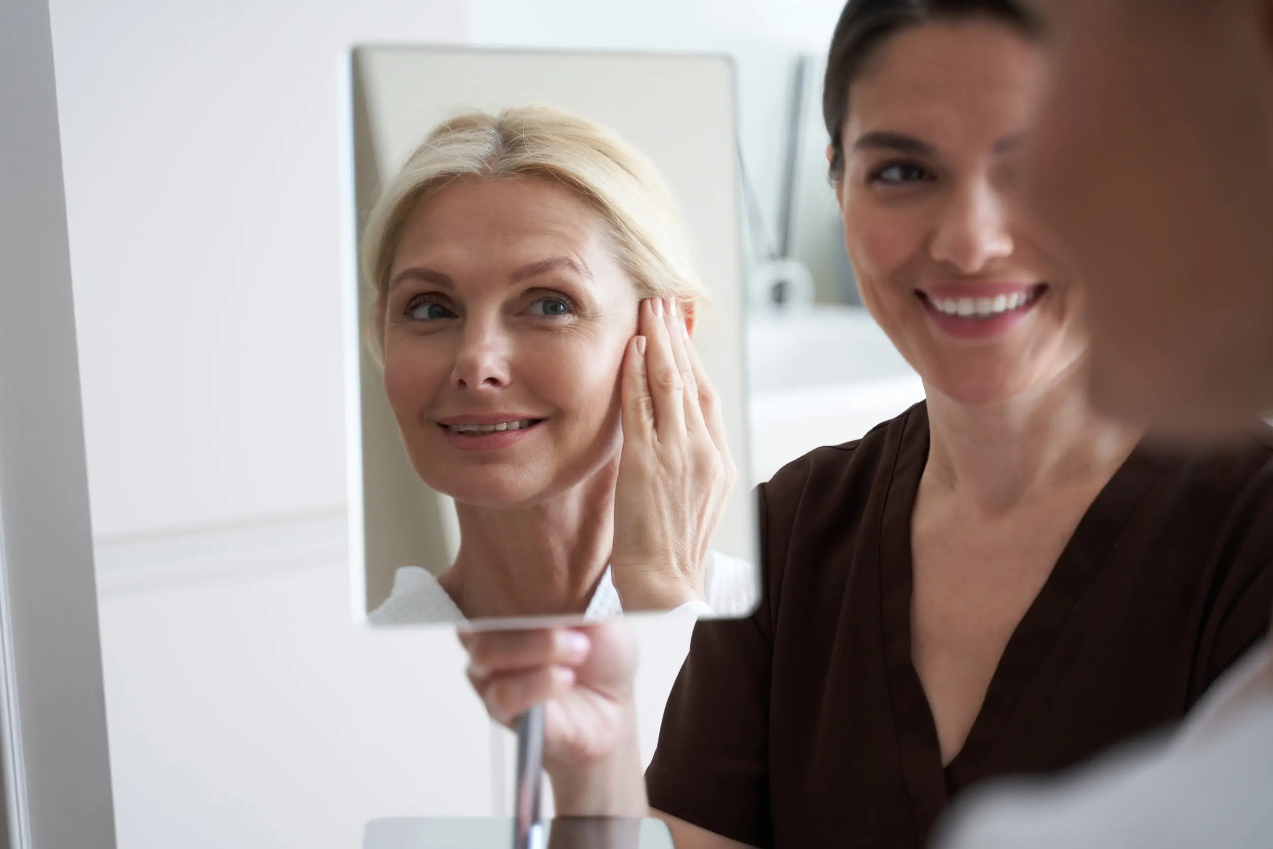 Two women smile at a mirror together, appearing pleased after facial surgery.