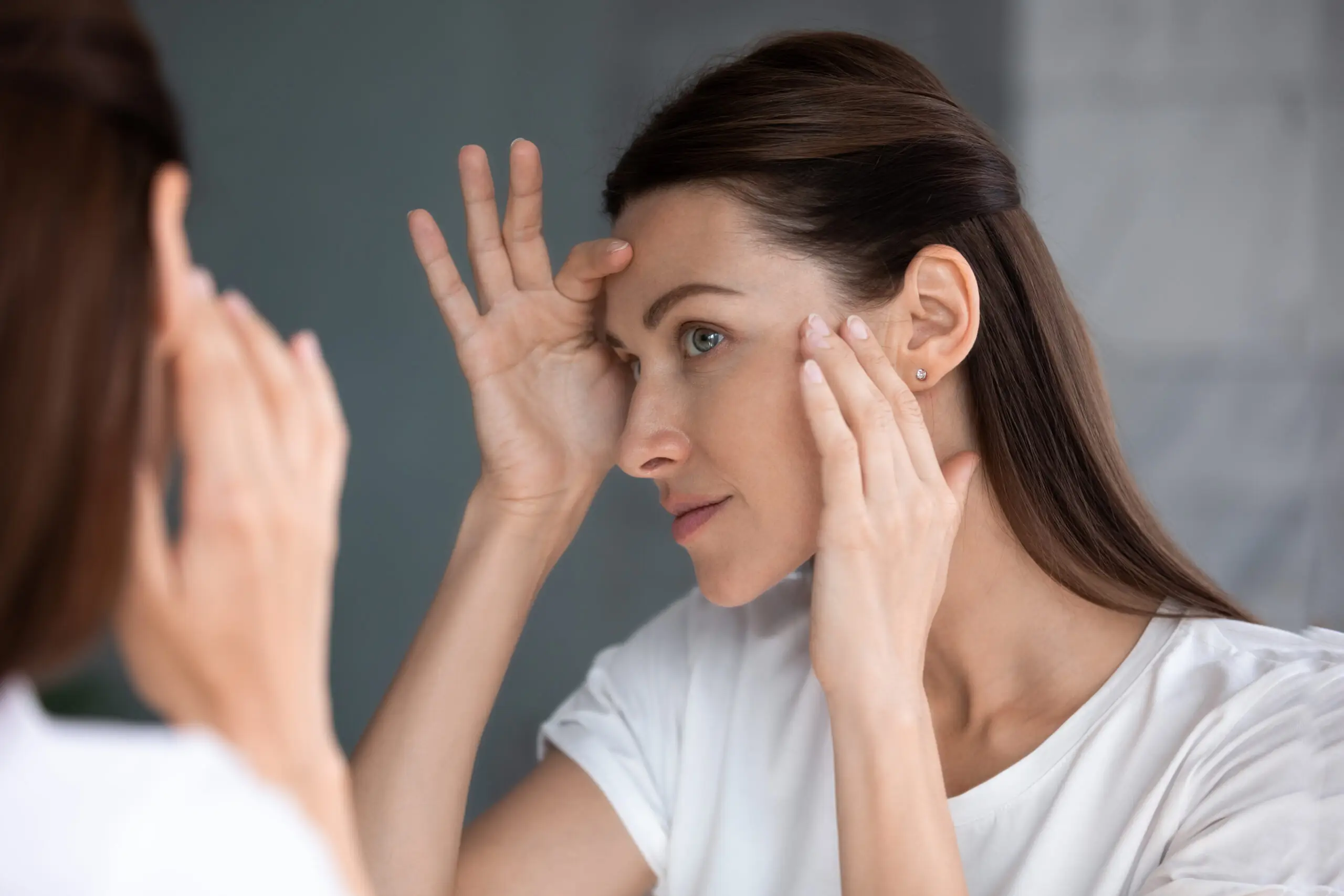 Woman in white shirt touching forehead and cheek while studying her reflection.