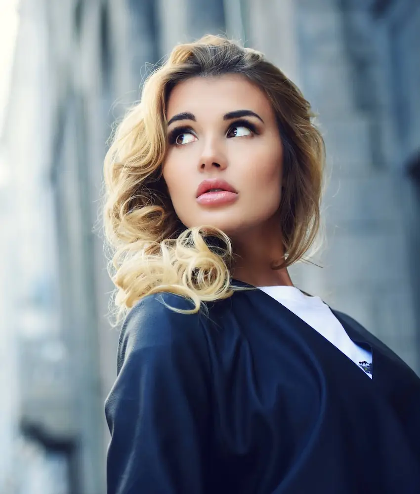 Woman with wavy blonde hair in dark jacket looks up outdoors near gray building.