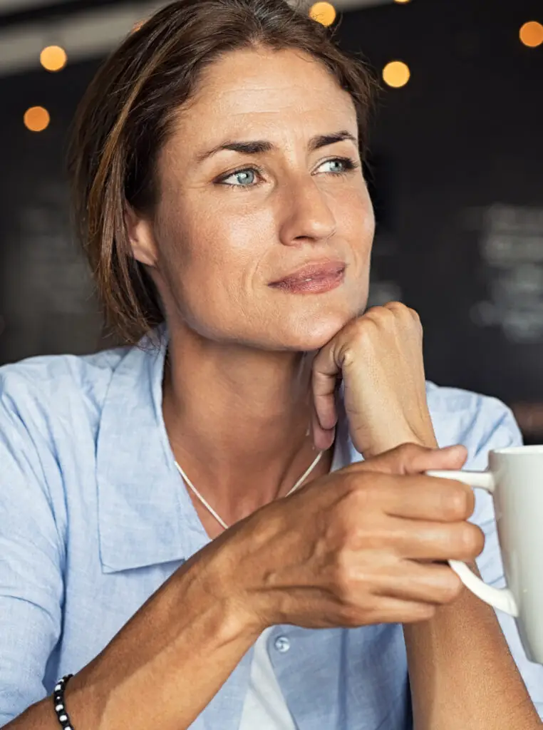 Woman with light eyes and brown hair holds mug, appearing thoughtful after facelift.