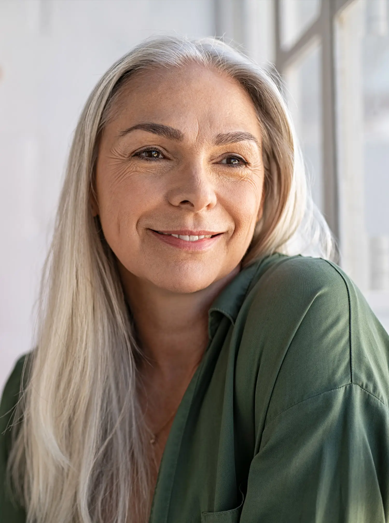 Older woman with long gray hair smiles by a sunny window at home.